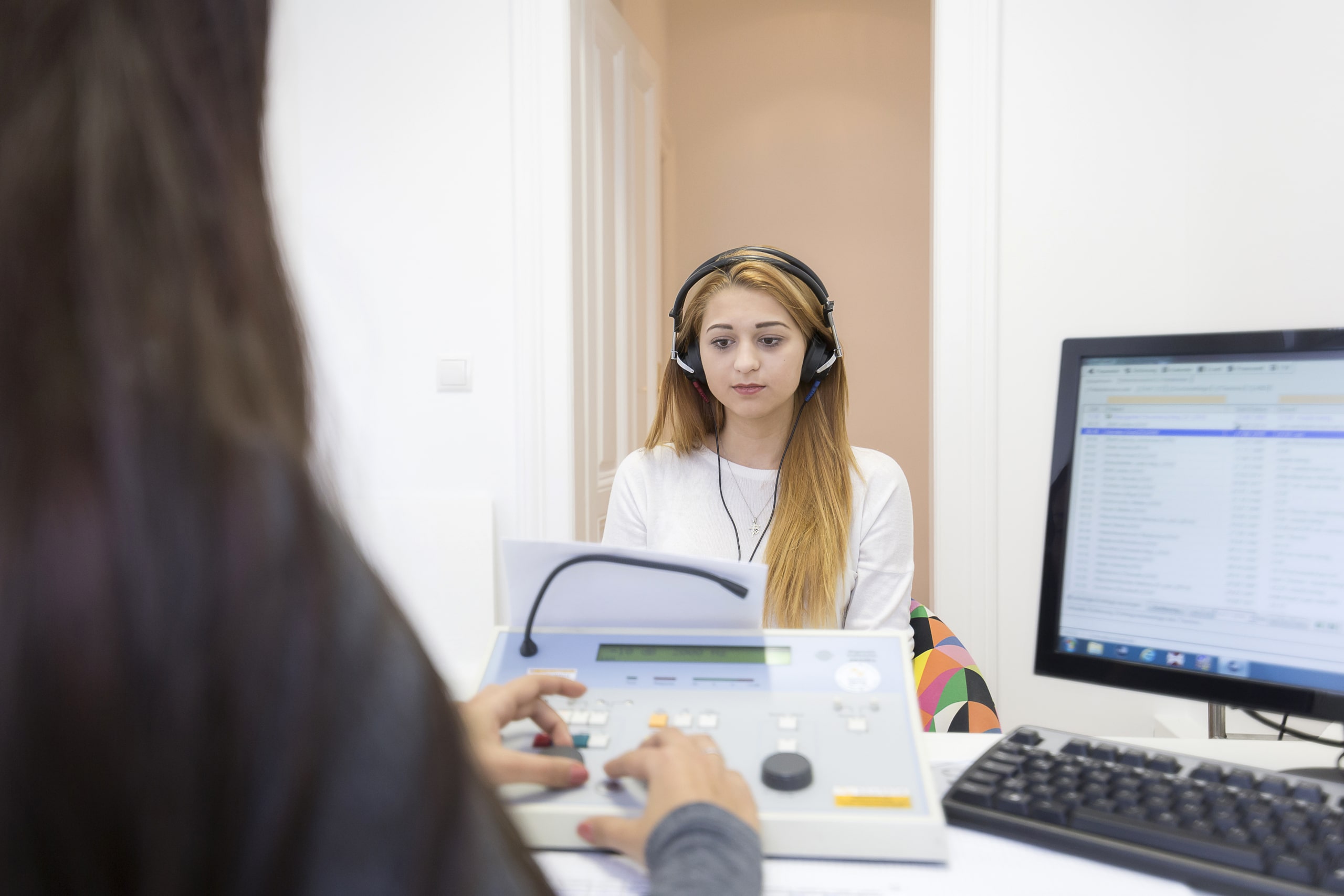 Frau mit Kopfhörern beim Hörtest im Hörtestraum, vor ihr sitzt eine Person an einem Audiometer und bedient das Gerät, daneben ein Computerbildschirm mit Testergebnissen